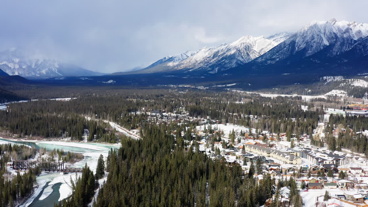 A serene drone flies in over Canmore and reveals a snowy forest, a frozen river, and the towering BANFF mountains beyond.