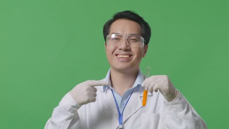 close up of asian man scientist smiling and pointing to orange liquid in the test tube in his hand while standing on the green screen background in the laboratory