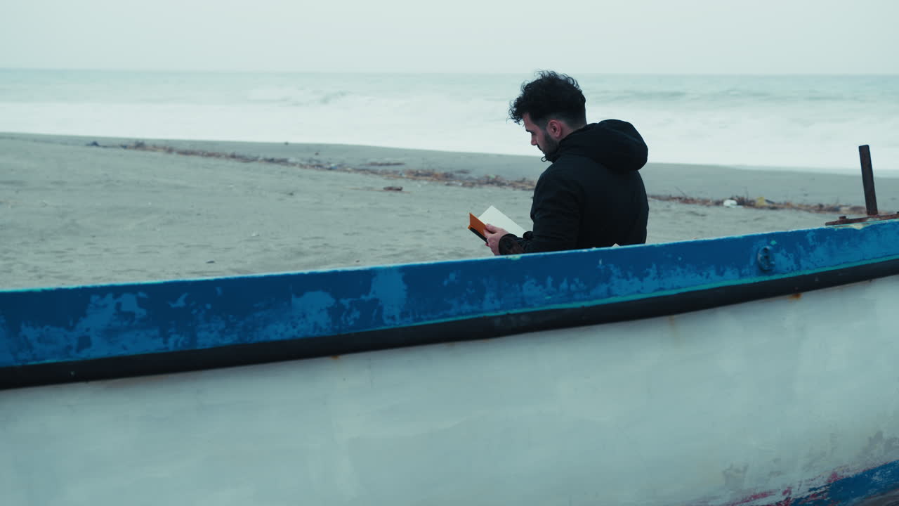 Man Reading A Book Sitting On The Beach In Winter Near A Boat