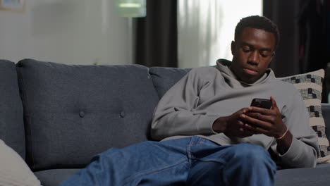 unhappy and depressed young man lying on sofa at home looking at mobile phone shaking his head