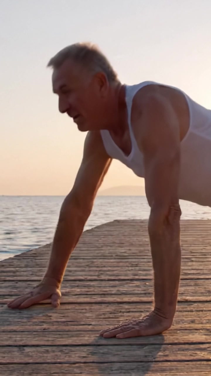 Uomo anziano impegnato in un esercizio di flessioni su un molo marittimo al tramonto, mostrando forza e determinazione nella routine di fitness all'aperto.