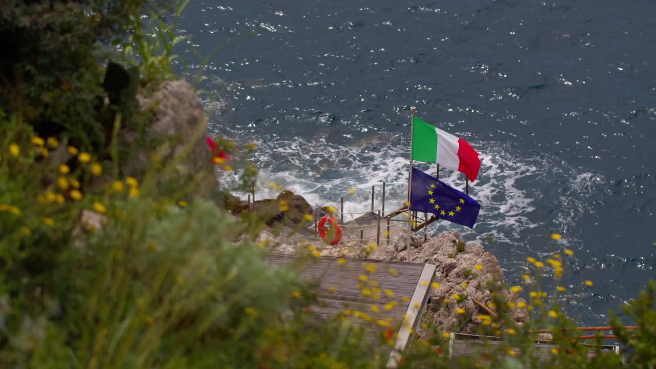 Bandiera italiana sopra la bandiera dell'UE che sventola su rocce costiere ventose a Taormina, Sicilia, Italia.