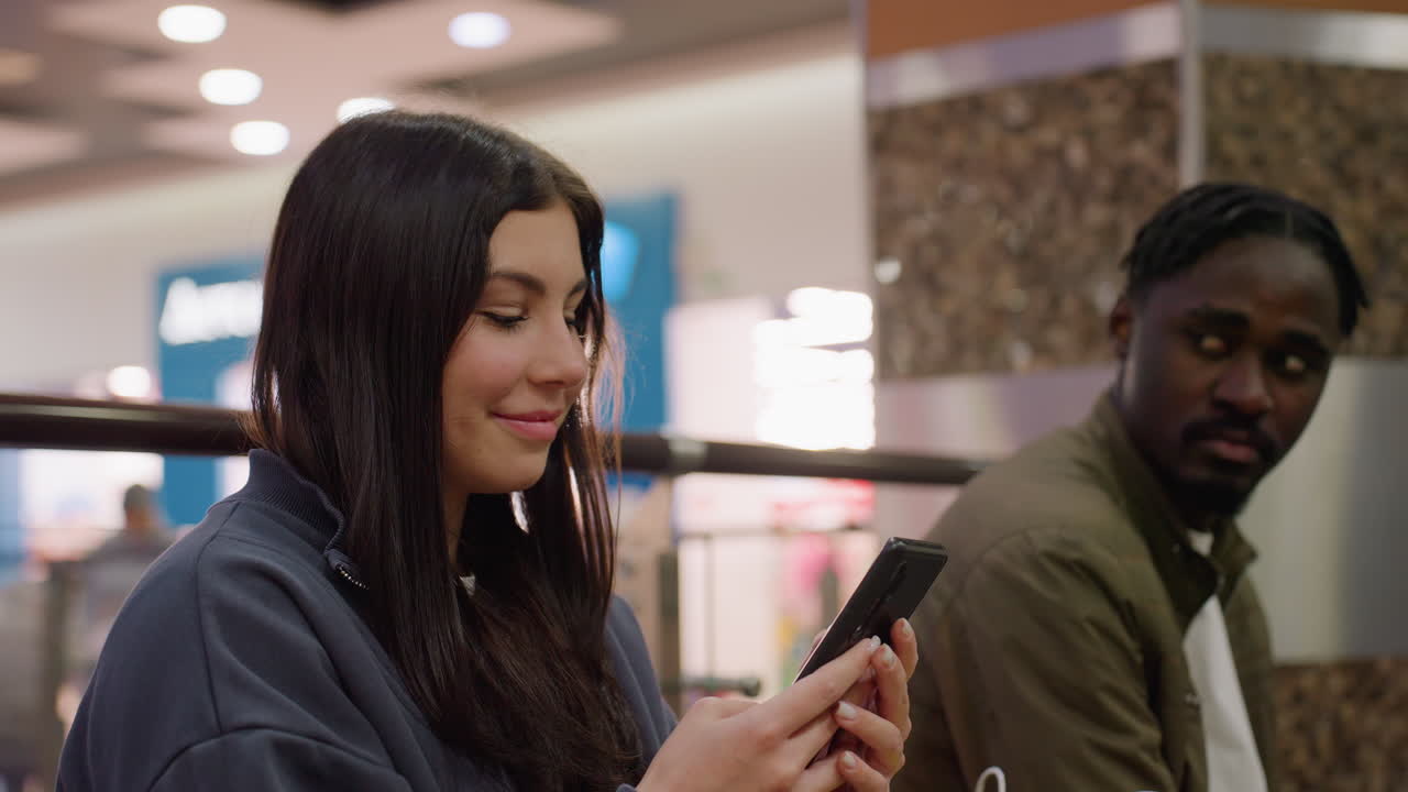 Smiling woman engaged with phone while husband turns away with visible frustration and sadness in blurred indoor mall setting, highlighting emotional disconnect and growing tension