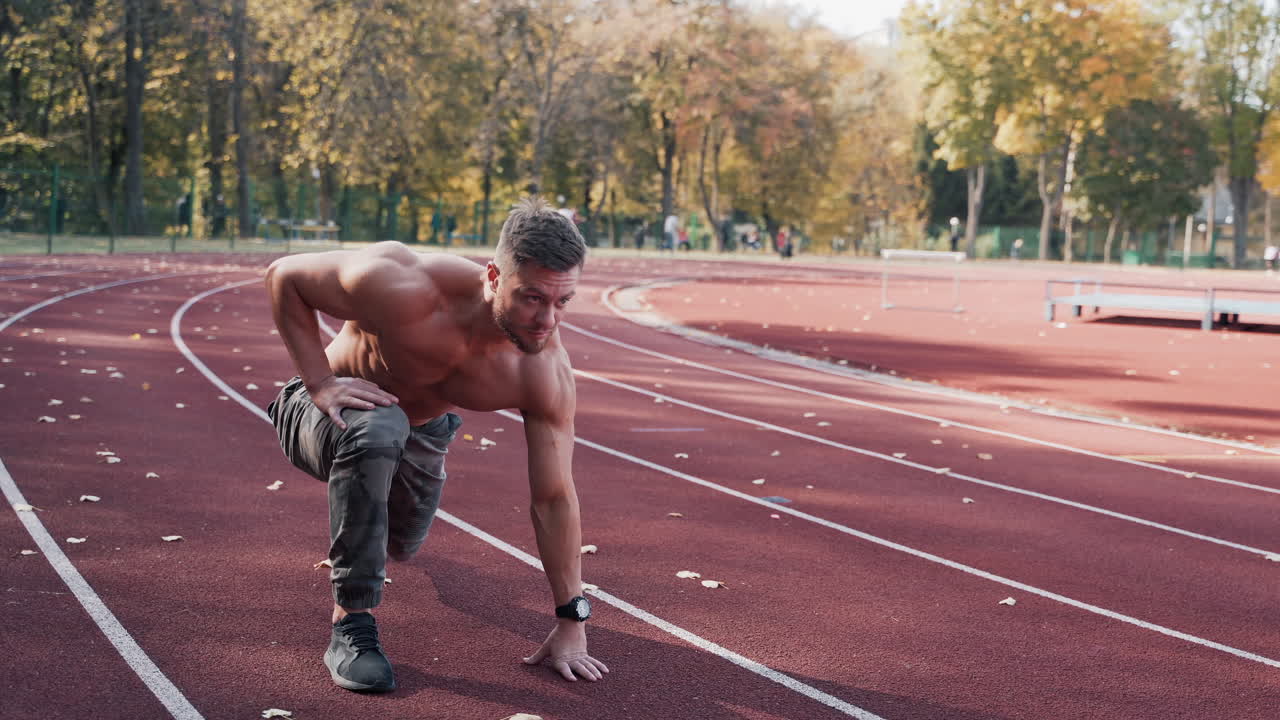 Shirtless athlete at the stadium. Strong muscular sportsman starts his running on the autumn background. Motion camera around.