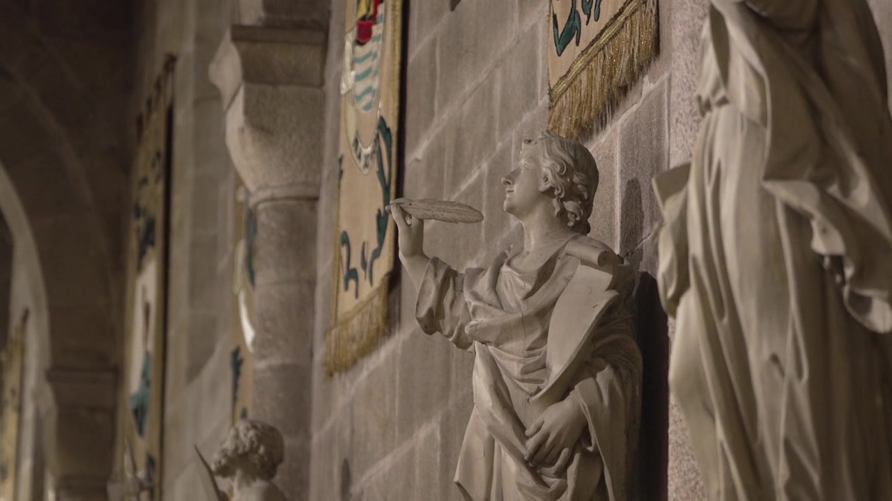 Primo piano di una statua di pietra che tiene in mano una penna d'oca, con stendardi sullo sfondo all'interno della Sé di Braga.