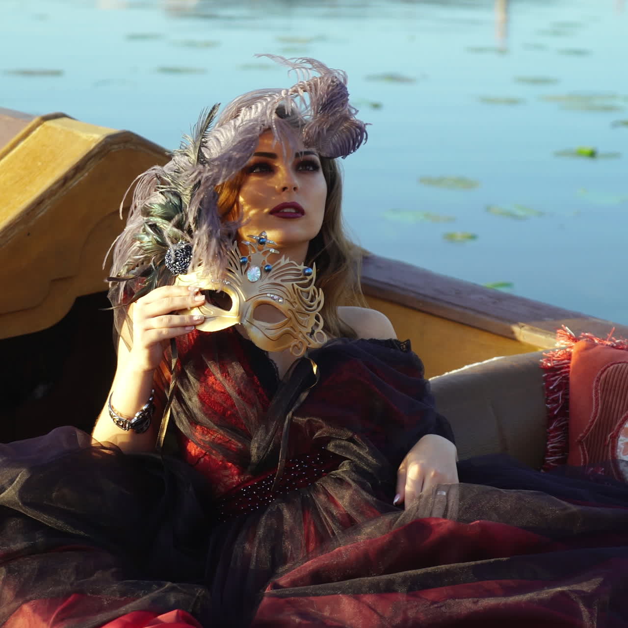 Romantic view of a beautiful woman in the evening dress lying in gondola over the water background. Attractive female holding carnival mask with a big feather outside in summer