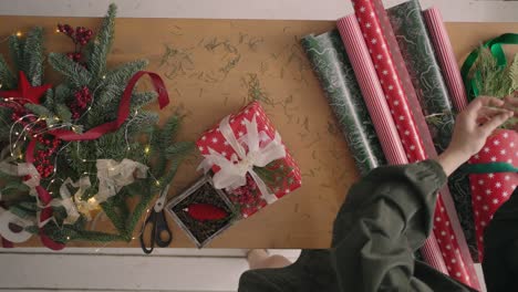 un plan de arriba hacia abajo. totalmente visible la mesa con las decoraciones. manos femeninas ponen y finalizan el regalo de navidad envuelto en papel de artesanía en una mesa de madera. cinta de vendaje