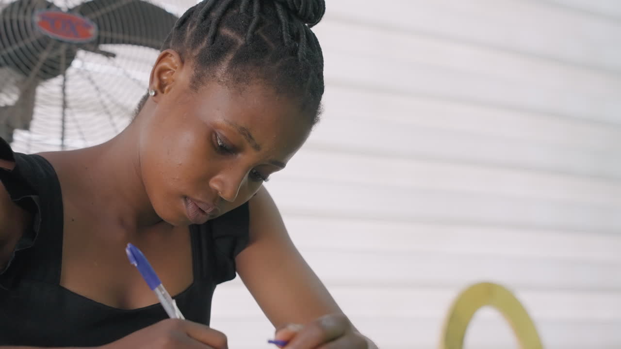 Woman calmly composing letter outdoors. Woman with braided hair concentrates on writing outdoors peacefully. Woman with braided hair peacefully drafts letter at table outdoors in sunlight