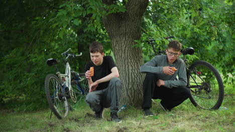 two young siblings enjoying snacks while squatting under a tree in a lush green forest, they are taking a break from cycling, with their bikes parked close by