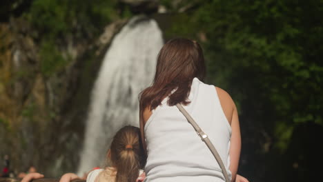 mother and little daughter enjoy waterfall view in wild nature park. loving girl leans on woman looking at water flow cascade at reserved area