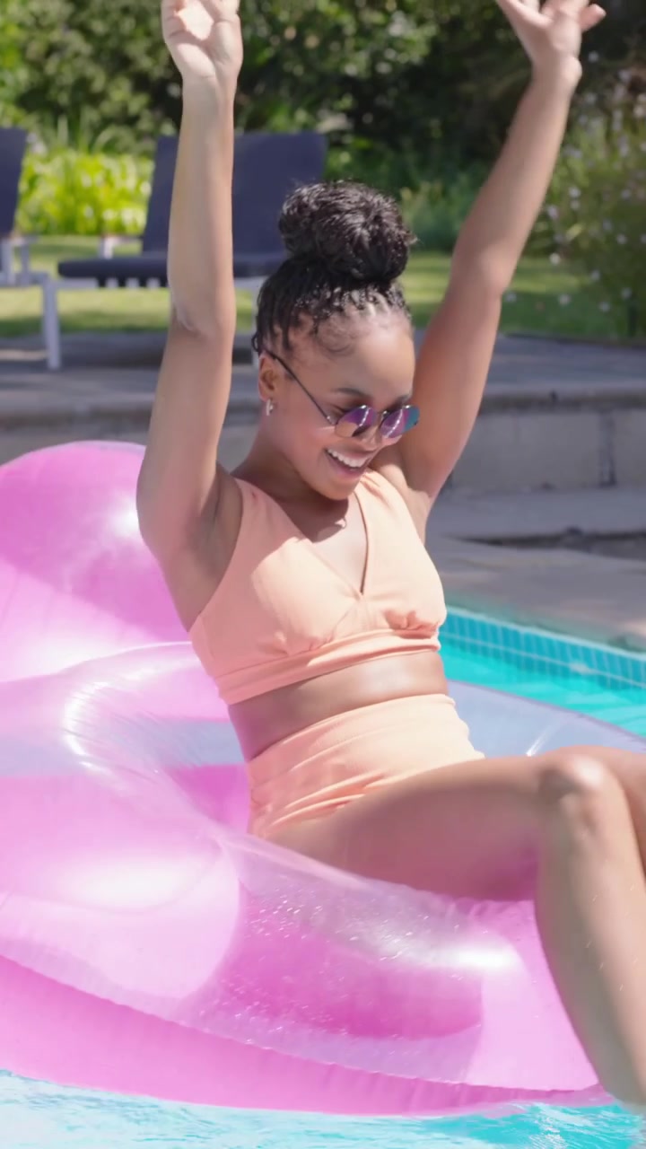Vertical video: Laughing woman in peach bikini climbing pink float at pool as man nudging playfully
