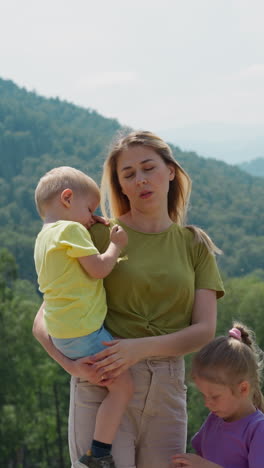 pensive tired woman tourist with little unhappy children son and daughter stands against large green hills on sunny summer day slow motion