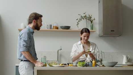 Mujer-Bonita-De-Pie-Preparando-Tostadas-De-Aguacate,-Esparciendo-Puré-De-Aguacate-En-Pan-Con-Un-Tenedor,-Mientras-Su-Marido-Habla-De-Algo-Interesante