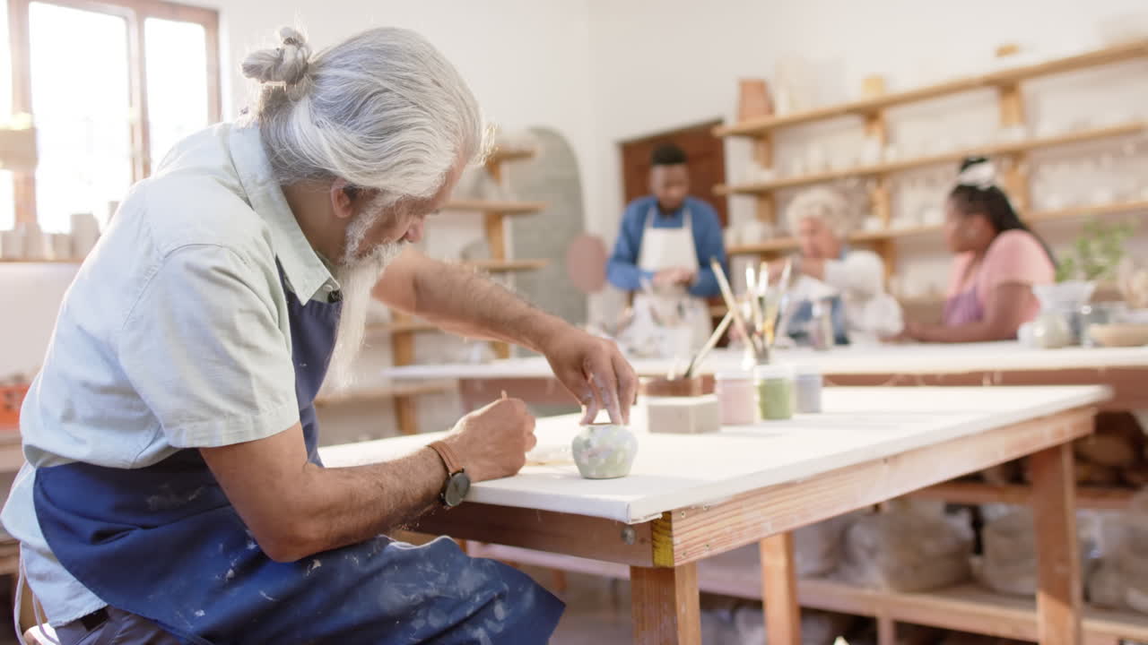 Focused biracial potter with long beard glazing clay jug in pottery studio, slow motion