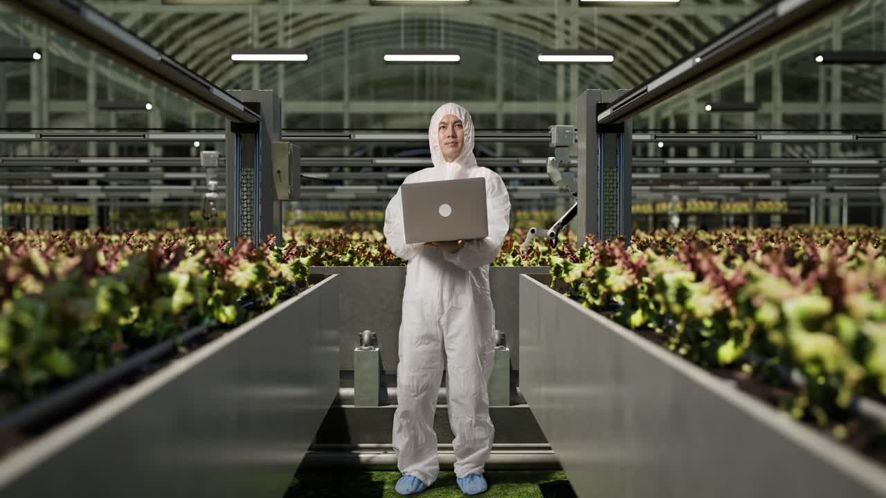 cuerpo lleno de investigador hombre asiático usando una computadora portátil y mirando a su alrededor mientras estaba de pie en el invernadero con agricultores robóticos inteligentes