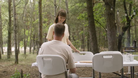 숲 속의 캠핑에서 남편과 아침 식사를 하기 위해 프라이팬을 테이블로 가져오는 행복한 여성