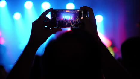 person capturing live music performance on smartphone, surrounded by vibrant stage lights, showcasing excitement and energy of concert atmosphere with audience in background
