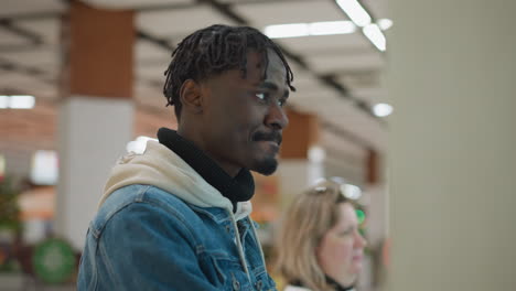 close-up of young man in denim jacket and hoodie, focused on self-service kiosk screen in food court. a woman is in the background, also interacting with nearby digital screen in busy mall environment