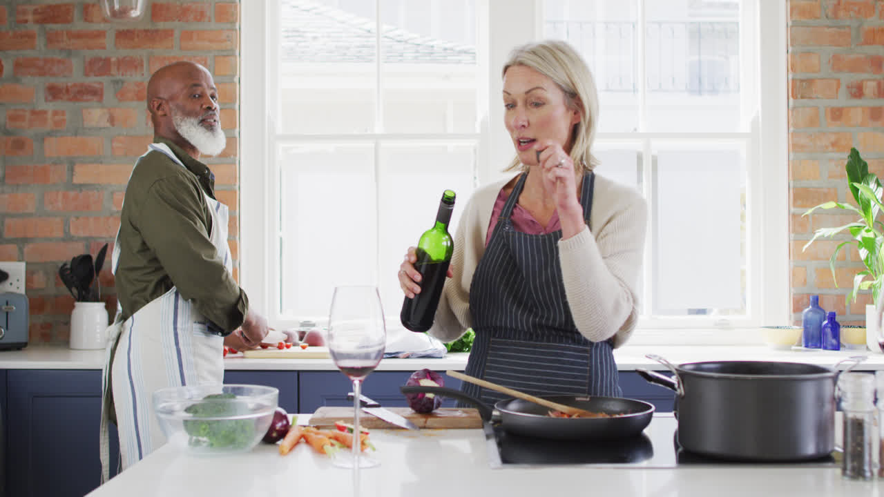 Eldre kvinne iført forkle heller vin i et glass på kjøkkenet hjemme.