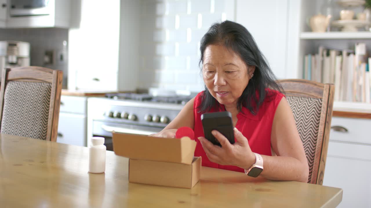 Senior Asian woman in red top opening package at kitchen checking smartphone and inspecting bottle