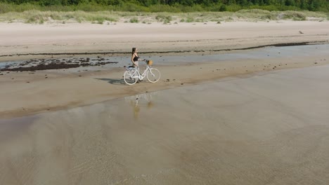 Vista-Aérea-Con-Una-Joven-De-Pelo-Largo-Parada-Con-Una-Bicicleta-En-La-Playa-De-Arena,-Día-Soleado,-Playa-De-Arena-Blanca,-Concepto-De-Estilo-De-Vida-Activo,-Amplio-Disparo-De-Drone-Ascendente-Moviéndose-Hacia-Atrás