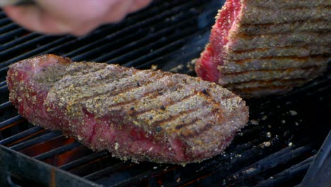 a pair of meat tongs lift a nearly cooked juicy rib eye steak on a grill and presses on it and another steak in slow motion