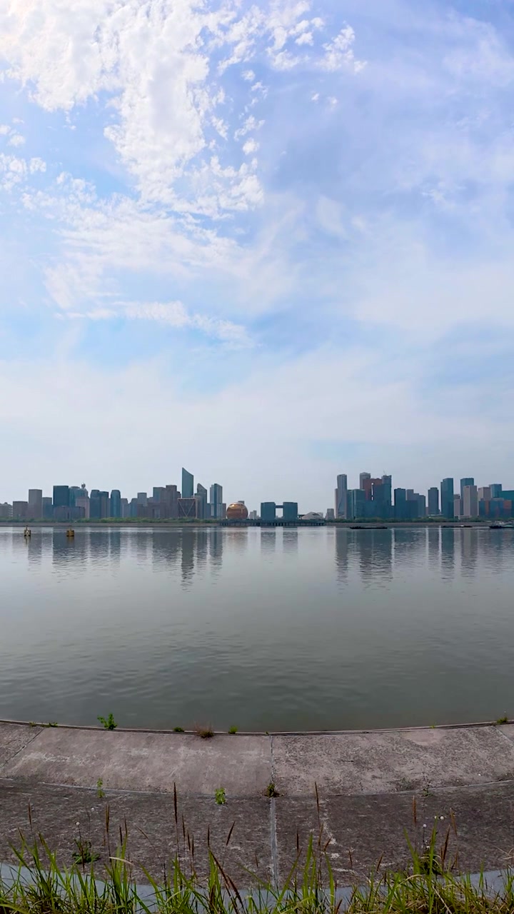 Vista vertical em time-lapse do horizonte de Hangzhou sobre o Rio Qiantang com reflexos calmos e arquitetura urbana moderna sob céus diurnos claros. UHD.