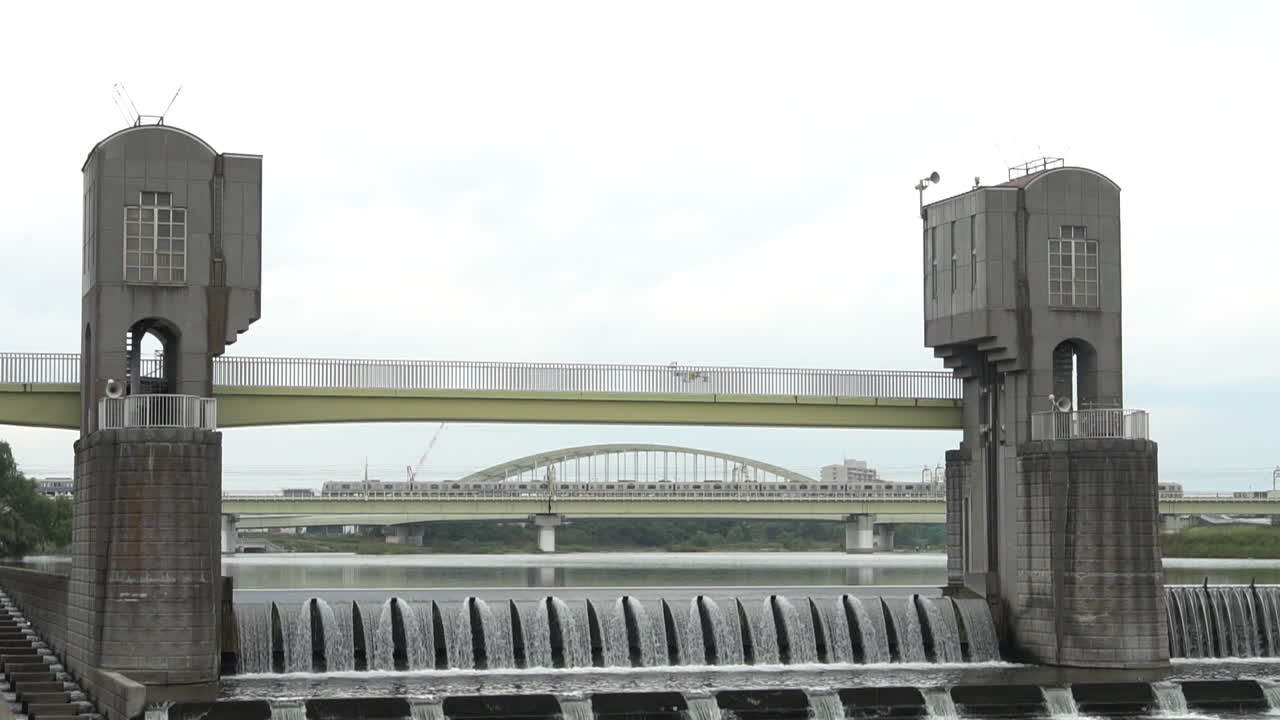 Water Flowing from Tama Ward Weir with Two Trains Intersecting on the Background. Wild Birds Can be seen from the area. It was taken on a Gloomy day of Autumn Season at Tama Ward, Kawasaki, Japan