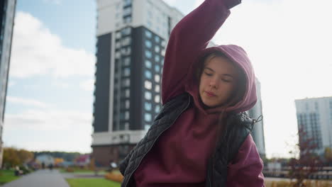 mindfulness coach in hoodie and quilted jacket stretches side muscles outdoors under bright sun against urban skyline with tall apartment buildings and autumn foliage on plaza