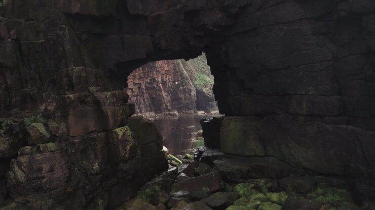 Aerial drone view moving through the Gatklettur sea arch with dark basalt walls rocky terrain and a natural opening revealing the sea beyond in Iceland