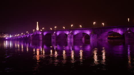 a beautiful arched stone bridge lit in purple, reflecting on a calm river at night
