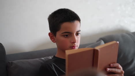 thoughtful kid with intense focus reading on couch flipping through book pages in calm indoor setting while partially visible lady sits nearby
