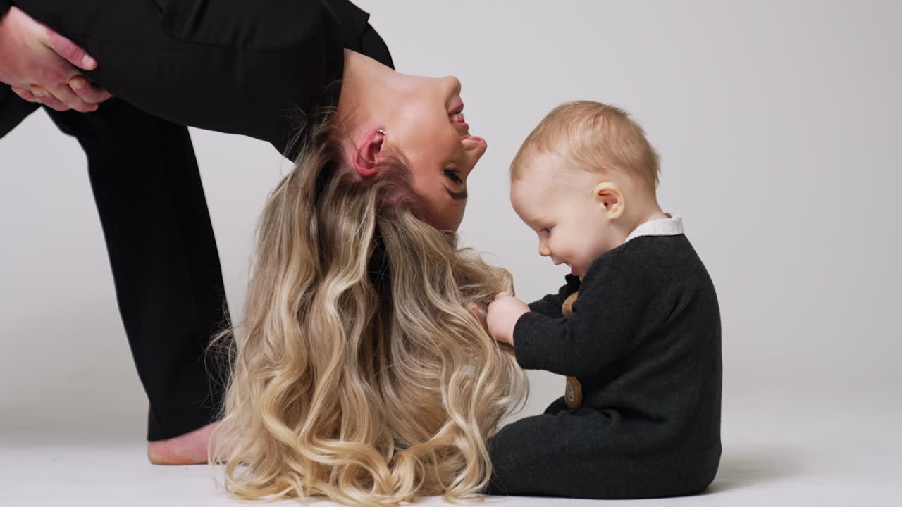 Baby sits on the floor touching long wavy hair of his mom. Mother kisses her cute child on the forehead.