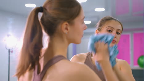 athletic woman in brown sportswear wipes large mirror with blue cloth in brightly lit fitness studio, mirror reflects background with gym equipment, pink window covers, and modern interior design