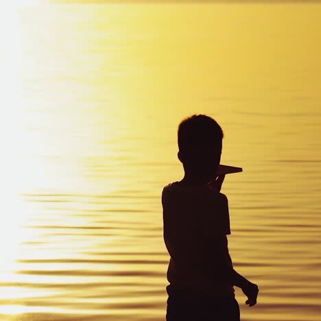 ein kleiner junge hält bei sonnenuntergang ein papierflugzeug in der hand. ein kind hebt seine hand zum himmel und spielt abends auf der straße mit origami