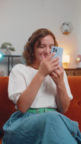 happy young woman sitting on sofa using smartphone celebrating success victory winning lottery