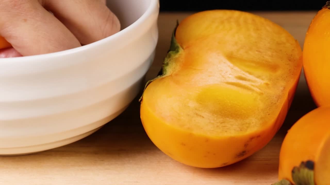 A hand reaches into a white bowl next to vibrant persimmons on a wooden surface.