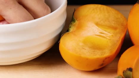 a hand reaches into a white bowl next to vibrant persimmons on a wooden surface.