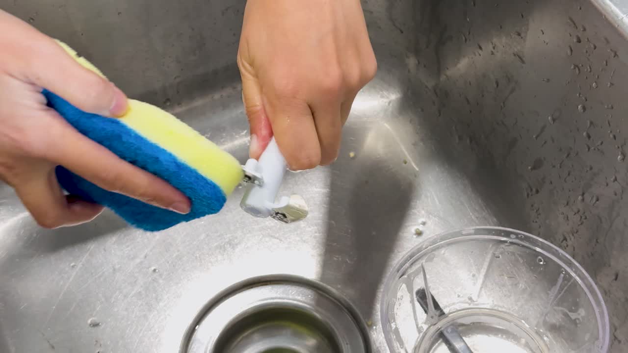 En person bruger en blå og gul svamp til at vaske en blenderkande og kniv i hånden i en køkkenvask af rustfrit stål under klar, jævn belysning.