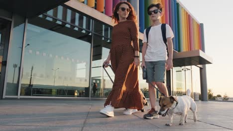 niño feliz, mujer con una maleta naranja en vestido largo y perro jack russell caminando a lo largo de la terminal del aeropuerto en lente llamaradas puesta de sol en verano en cámara lenta. familia va de viaje. viaje. turismo