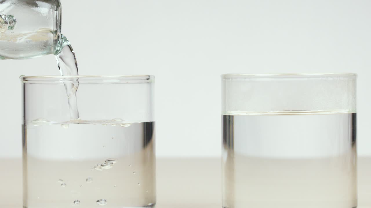 Clear glass pitcher pours cold water into empty glass, neutral background, bright, even lighting
