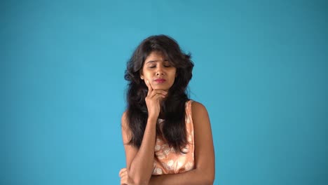 A-young-Indian-girl-in-orange-frock-wearing-giving-sad-thinking-expression-standing-in-an-isolated-blue-background