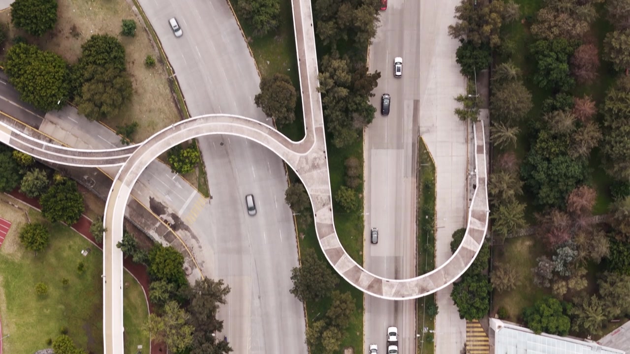 Aerial drone view of a wide, tree-lined boulevard with a curved S-shaped pedestrian bridge in Puebla, Mexico