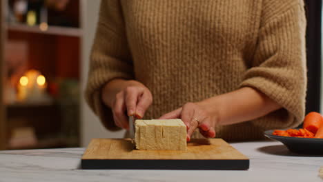 close up of woman at home in kitchen preparing healthy vegetarian or vegan meal slicing tofu on board with knife 1