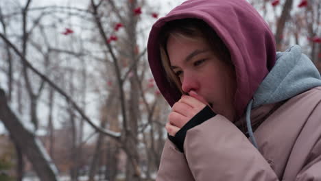 side view of a woman feeling cold as she blows warm air into her hands and adjusts her hoodie, she is bundled up in a thick jacket, standing amid bare trees with a snowy winter background