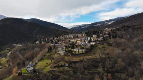 Antena:-Pequeño-Pueblo-De-Montaña-En-Un-Paisaje-Montañoso-Con-Montañas-Cubiertas-De-Nieve-En-El-Fondo,-Cielo-Azul-Y-Nubes