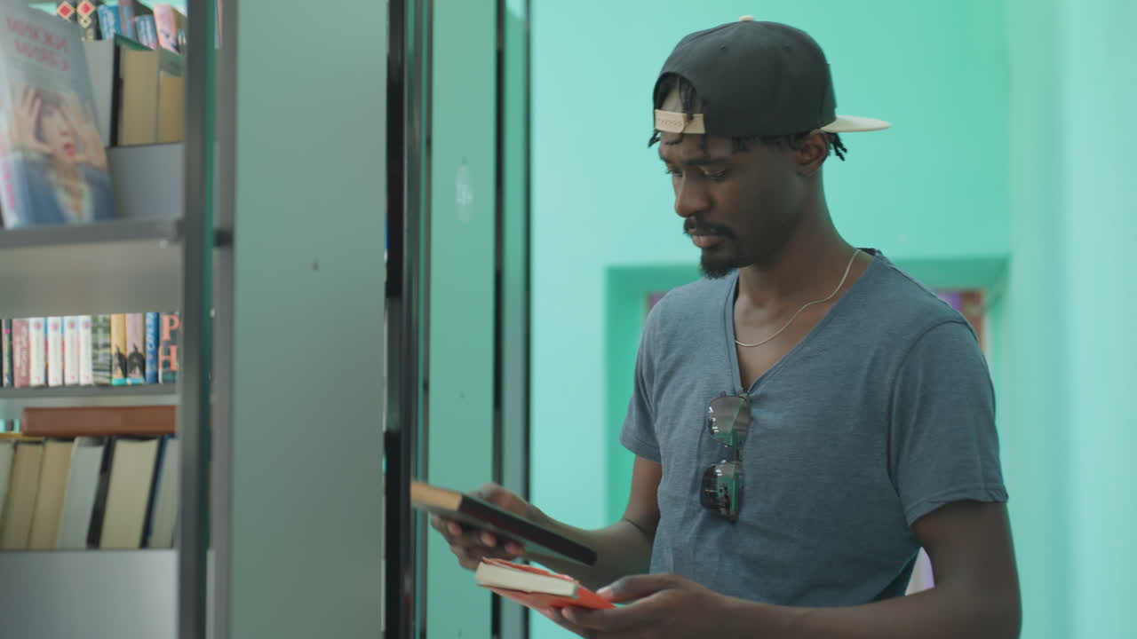 Student wearing cap and casual shirt reaches toward bookshelf, attentively examining book cover, with sunglasses hanging from shirt and teal walls contrasting purple hallway in background