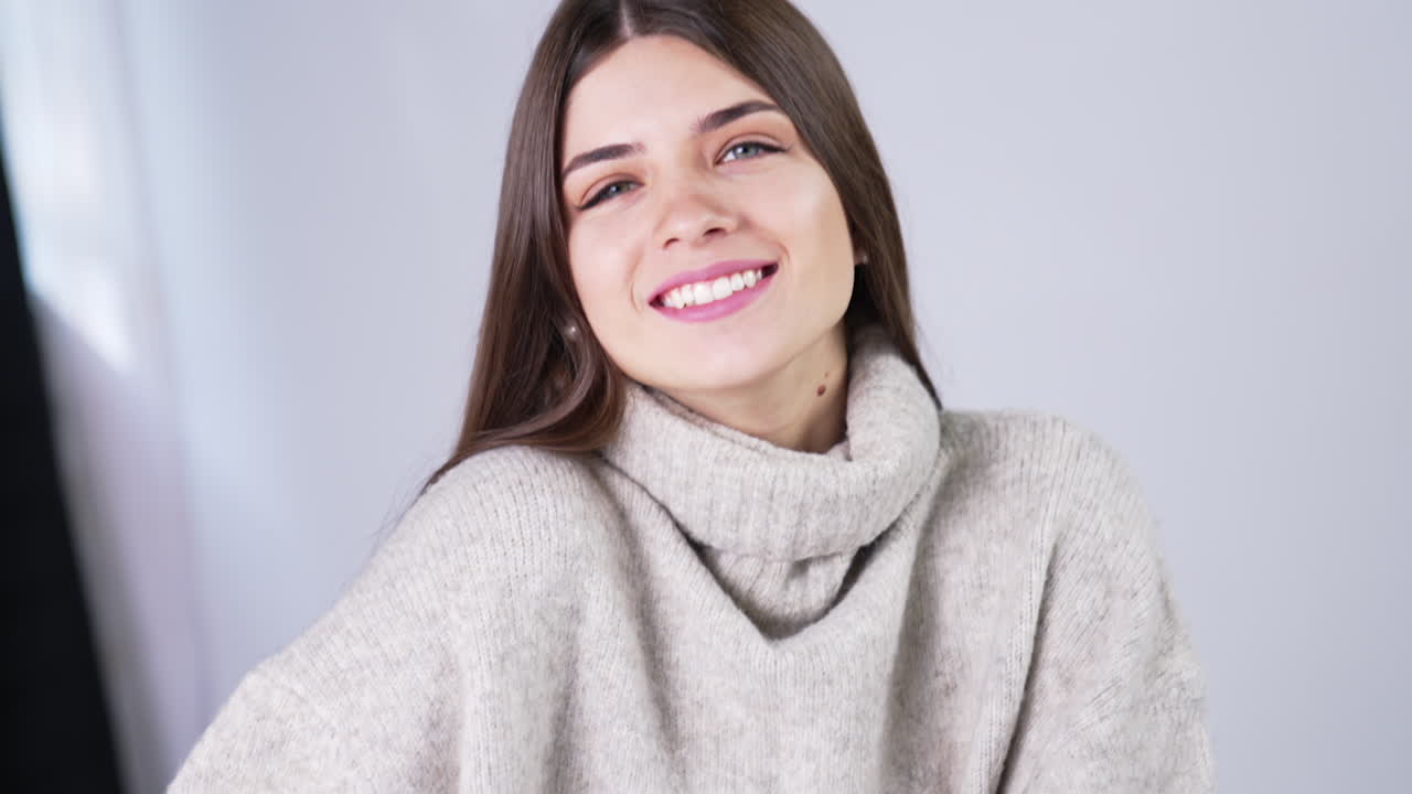 Cheerful smiling beautifully and touching her long dark hair. Female model wearing warm grey sweater posing in studio. Close up.
