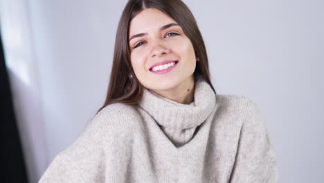cheerful smiling beautifully and touching her long dark hair. female model wearing warm grey sweater posing in studio. close up.