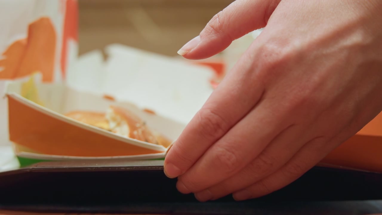 Lowangle hand slides tray under table, cinematic underside view of takeout tray being retrieved, neon reflections and patterned floor visible, careful fingers nudging cardboard box and cup, shallow